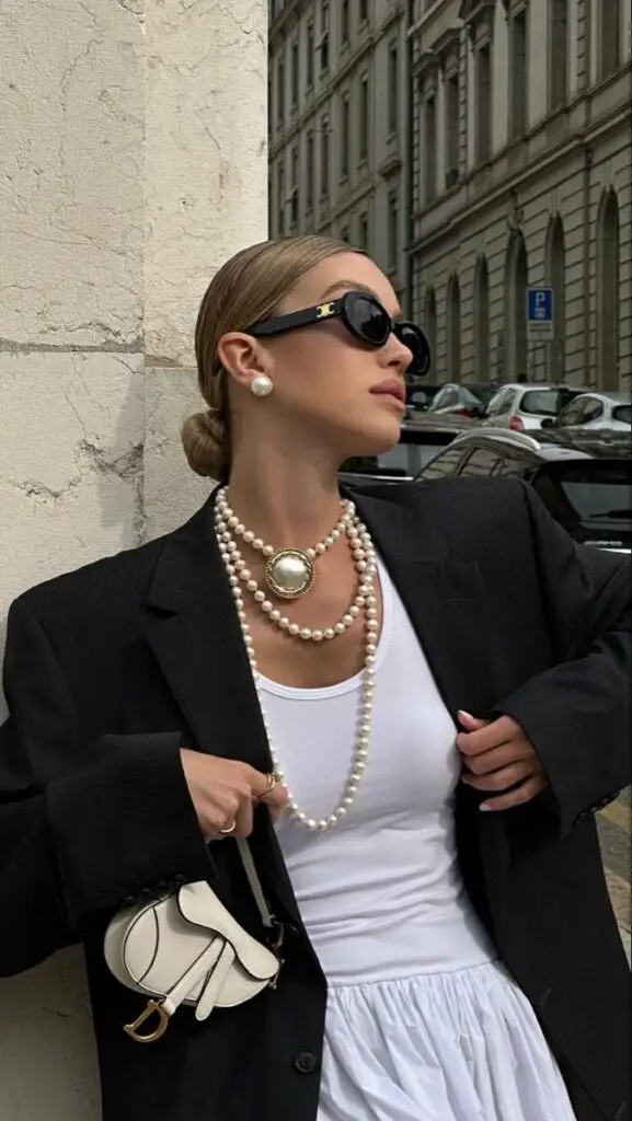 Woman in chic outfit with pearls, black blazer, white top, sunglasses, posing against urban backdrop.
