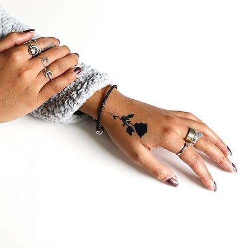 Person with rose tattoo on wrist, wearing silver rings and a gray sweater on a white background.
