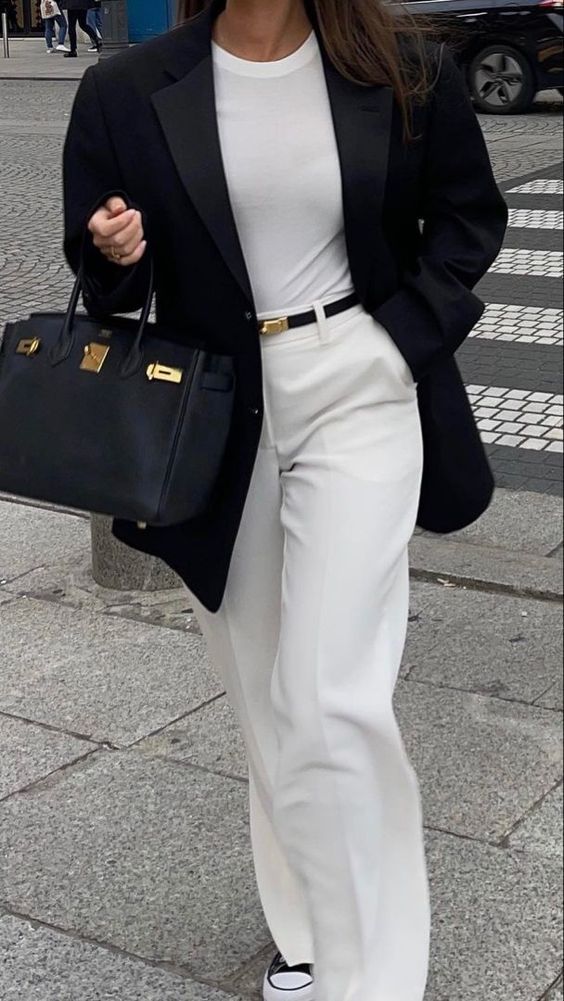 Stylish woman in white pants and black blazer holding a designer handbag, walking on a city street.