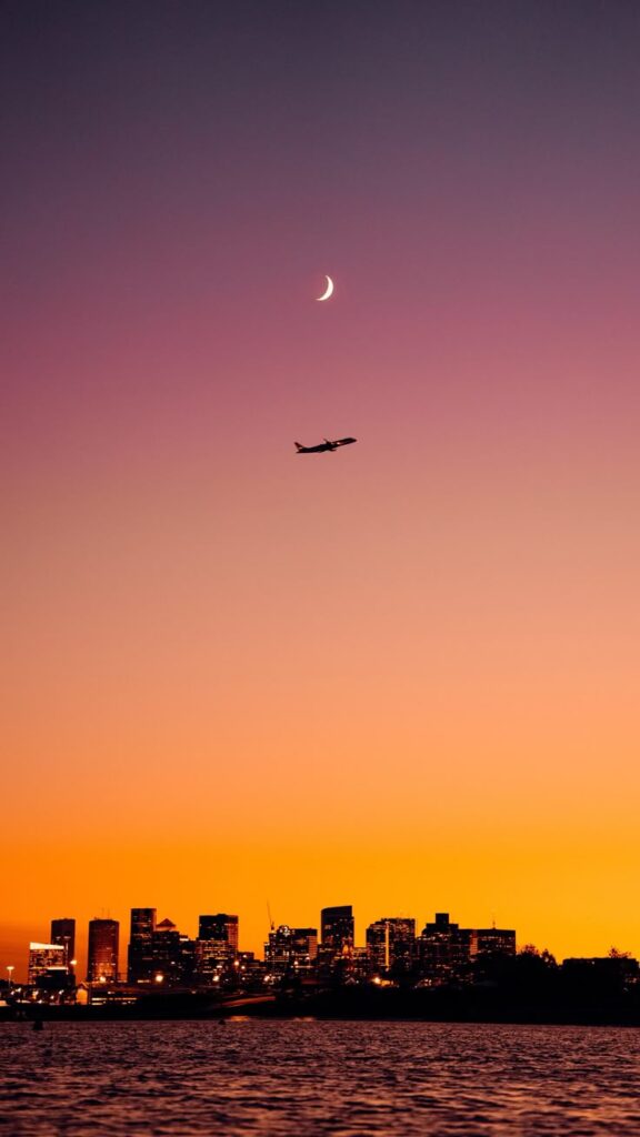 Airplane over city skyline at sunset, crescent moon in colorful sky, reflecting tranquility and travel possibilities.