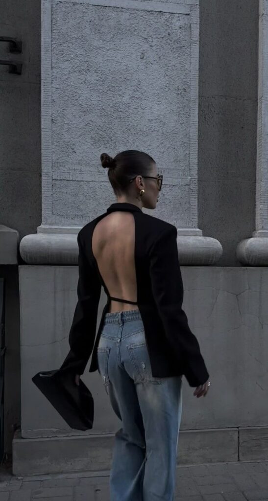 Fashionable woman in open-back blazer and jeans walking outside, holding handbag, showcasing urban style.