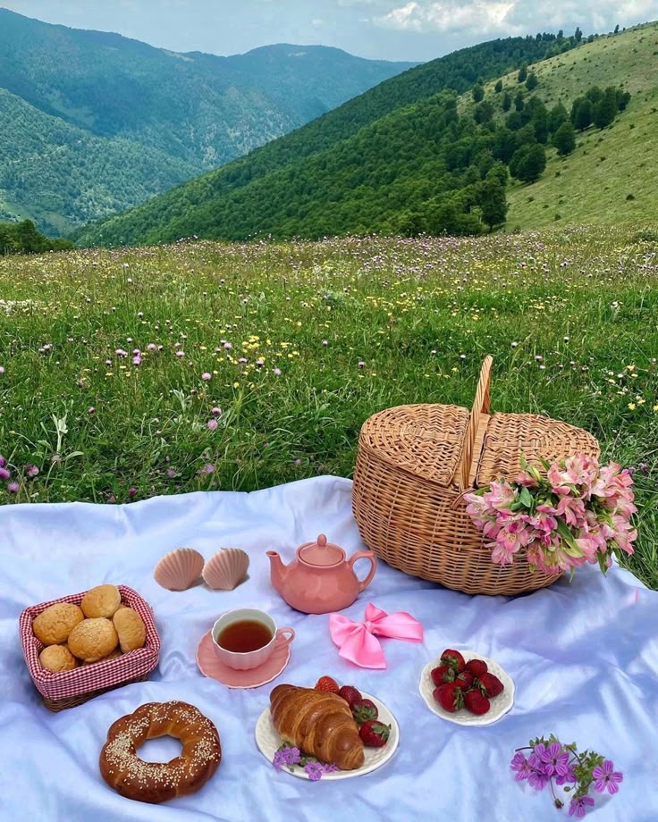 Picnic setup with pastries, tea, and fruits on a blanket in a scenic mountain meadow, wicker basket and flowers.