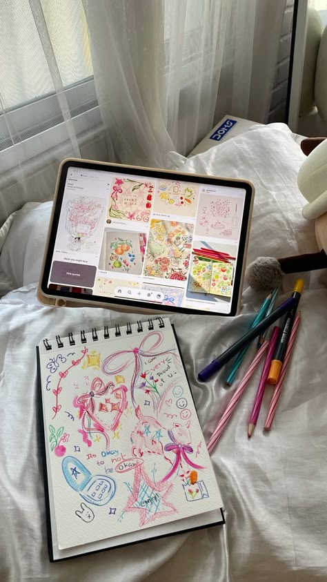 Tablet displaying colorful art ideas next to a sketchbook with similar creative doodles and pens on a bed.