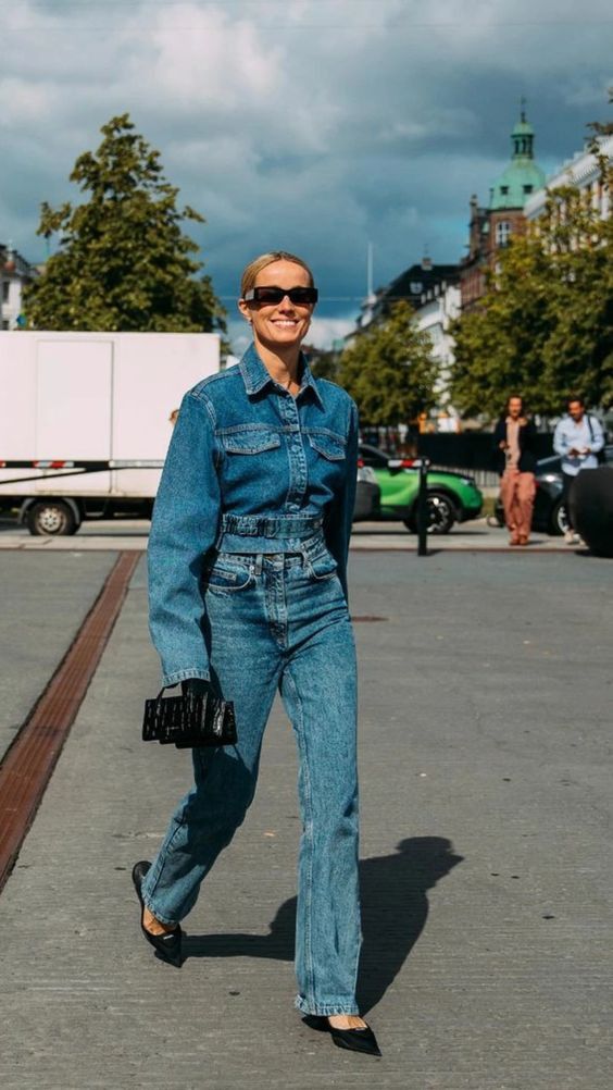 Fashionable woman in denim jacket and jeans walking outside with handbag, under a cloudy sky.