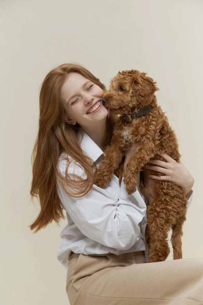 Smiling woman holding a curly brown dog that licks her cheek, capturing a joyful moment together.