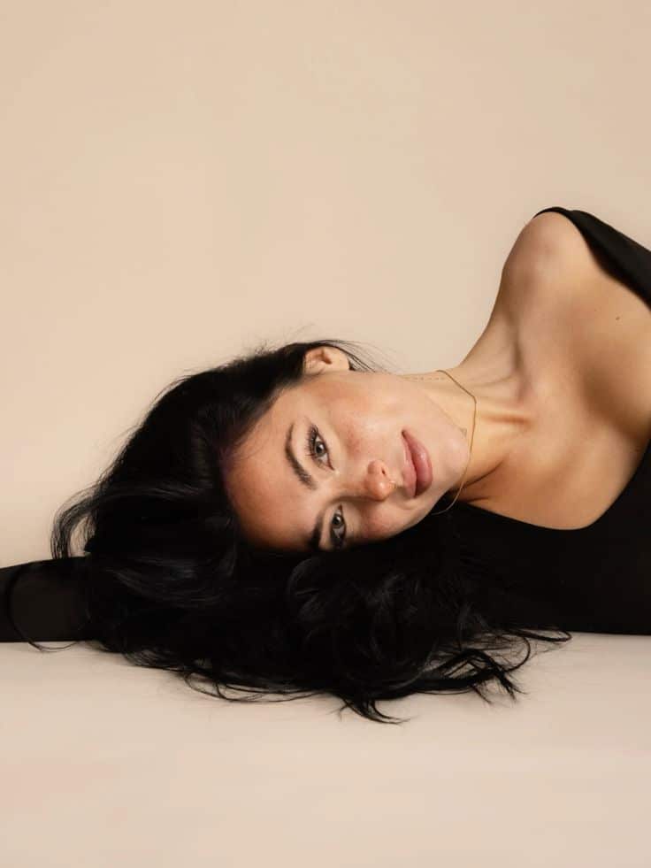 Woman lies on a beige background with black hair cascading, wearing a black top and gold necklace.