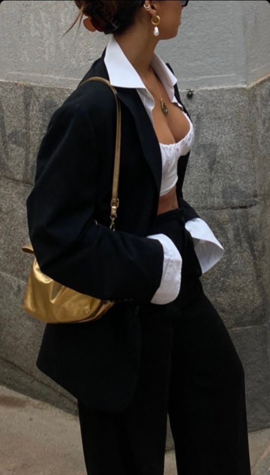 Stylish woman in black blazer with gold purse, white crop top, and pearl earrings on city street. Fashionable outfit.
