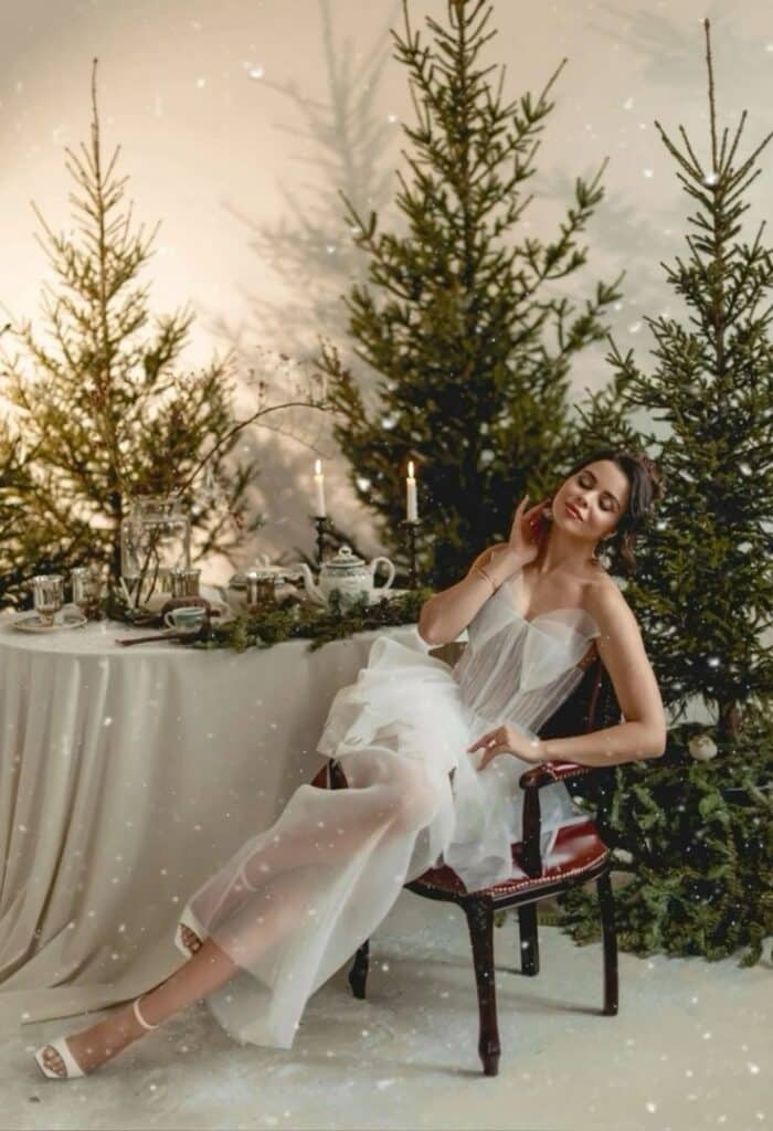 Elegant woman in white dress by a festive tablescape, surrounded by evergreen trees and candlelight.
