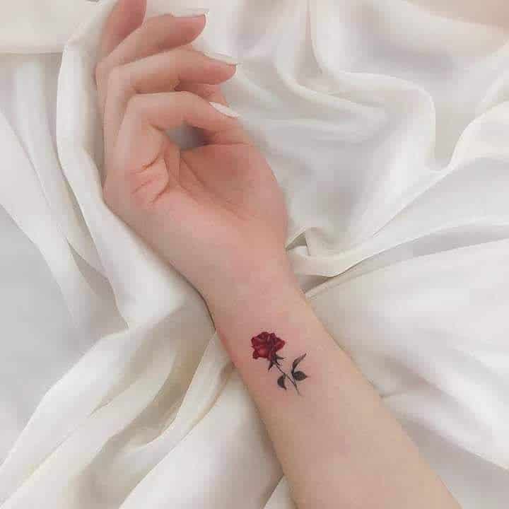 Delicate red rose tattoo on wrist against soft white fabric background, showcasing minimalist floral design.