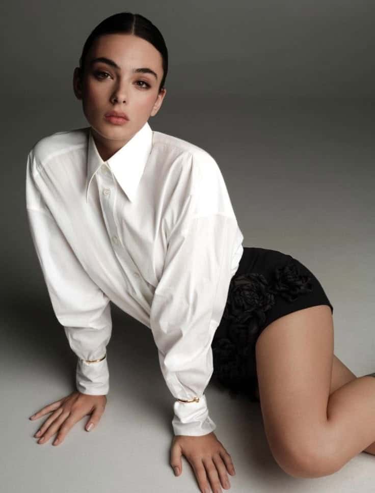 Woman in white shirt and black shorts kneeling on floor, looking forward. Fashion photoshoot with minimalist backdrop.