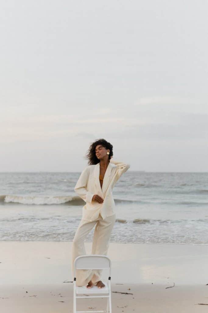 Woman in beige suit standing on the beach with ocean waves in the background.