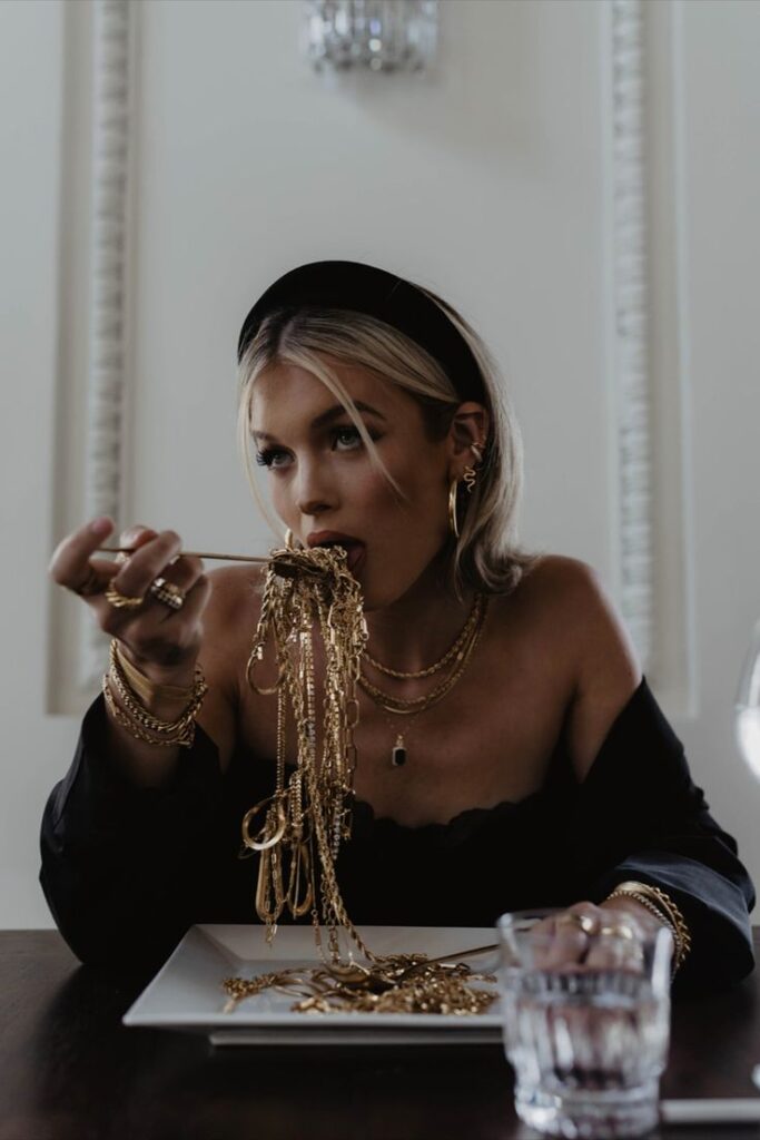 Woman wearing jewelry, playfully eating gold chains like spaghetti in elegant setting. Glamorous and artistic concept.