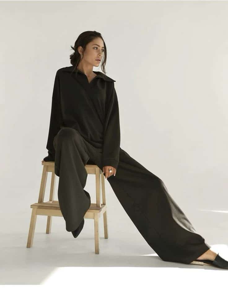 Person in oversized black outfit sitting on a wooden chair in minimalistic setting.
