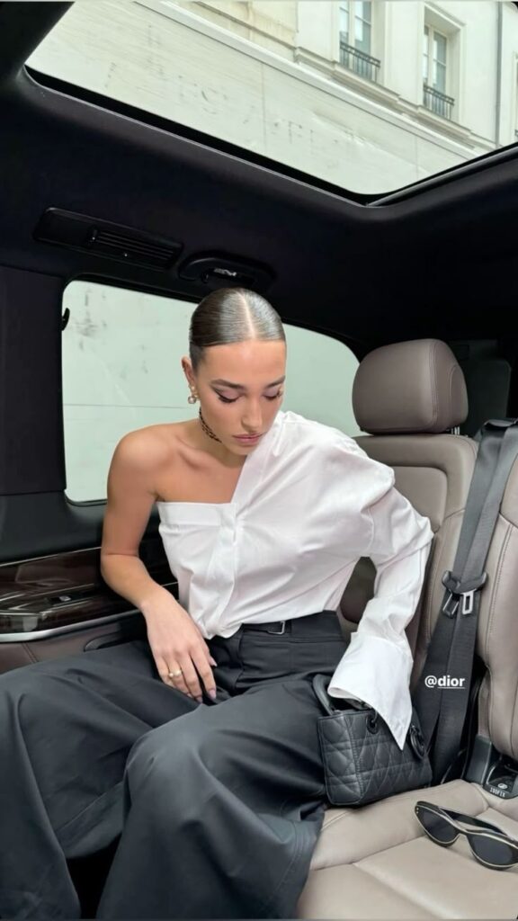 Elegant woman in a car, wearing a chic white blouse and black pants, with a quilted bag and sunglasses by her side.