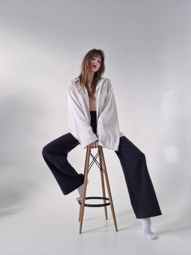 Person in relaxed pose on stool wearing white shirt and black pants, minimalistic studio background.