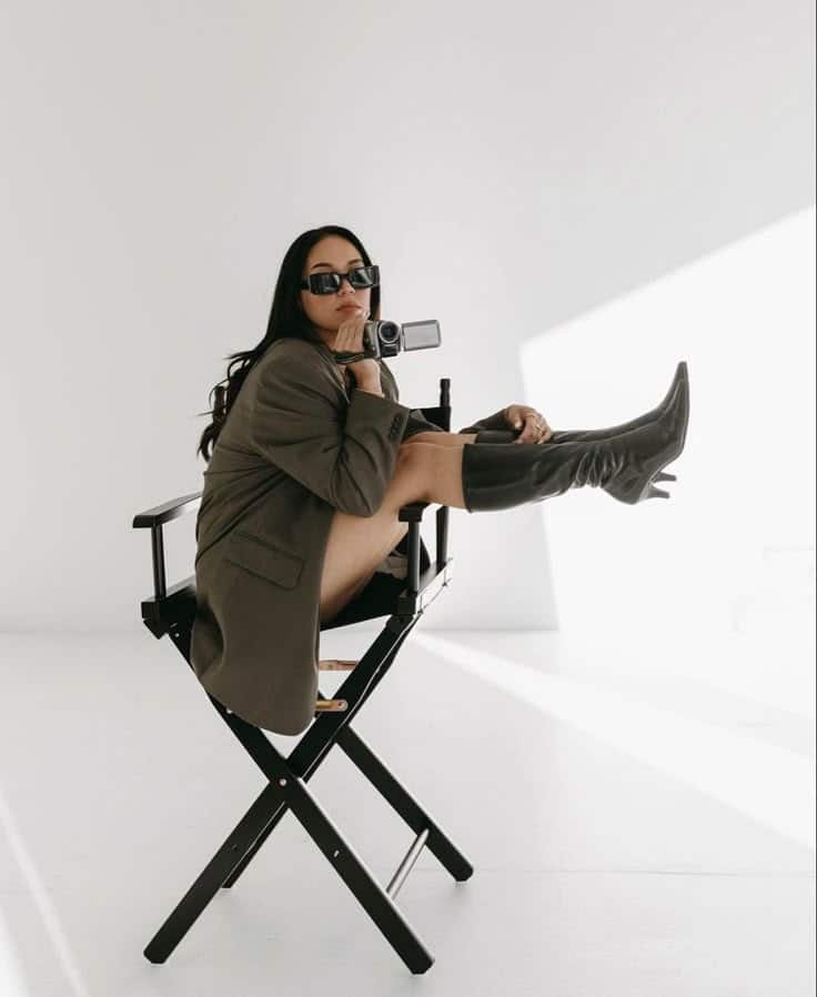 Woman in black boots and sunglasses sitting stylishly on a director's chair, posing with a vintage camera.