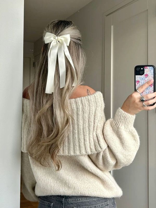 Blonde hair with a white bow, wearing an off-shoulder sweater, taking a mirror selfie with a floral phone case.