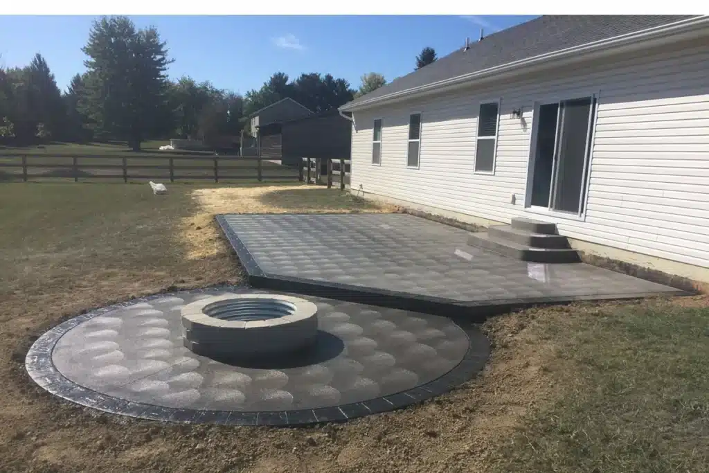 Backyard with gray patio extension and a circular fire pit next to house with sliding doors.