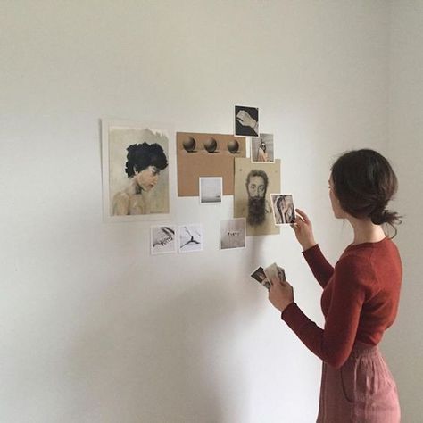Woman arranging art and photos on a minimalist wall gallery, wearing a red top and pink skirt, creating a personal display.