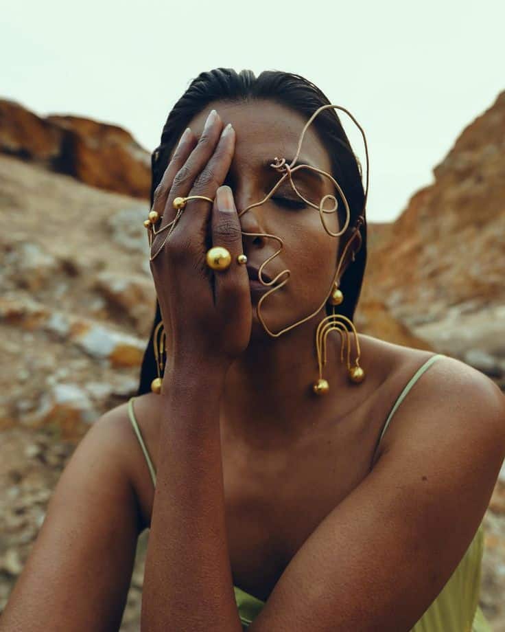 Woman wearing artistic gold jewelry and green outfit in desert landscape, with hand covering face. Elegant and stylish.