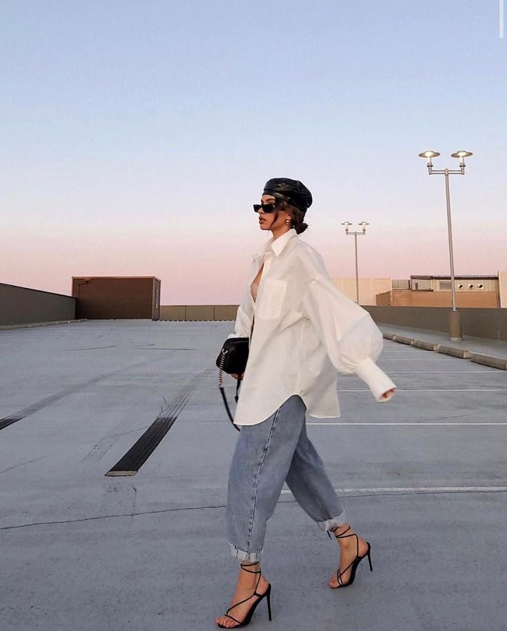 Fashionable woman in oversized shirt and jeans walking on rooftop at sunset, wearing heels and sunglasses.