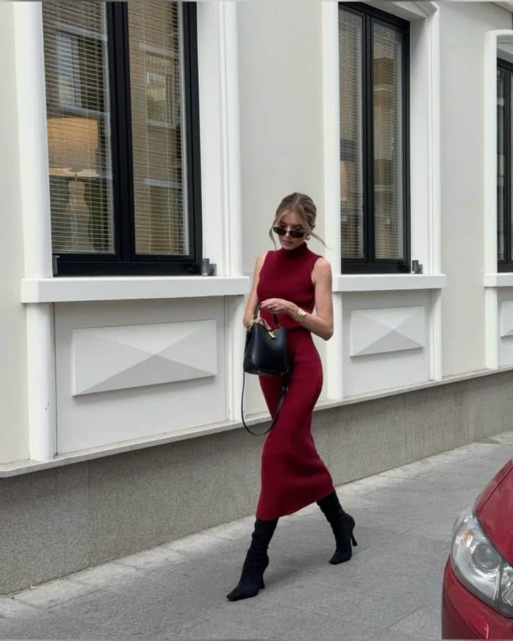 Woman in elegant red dress and black boots walking on city street, holding a black handbag near a modern building.