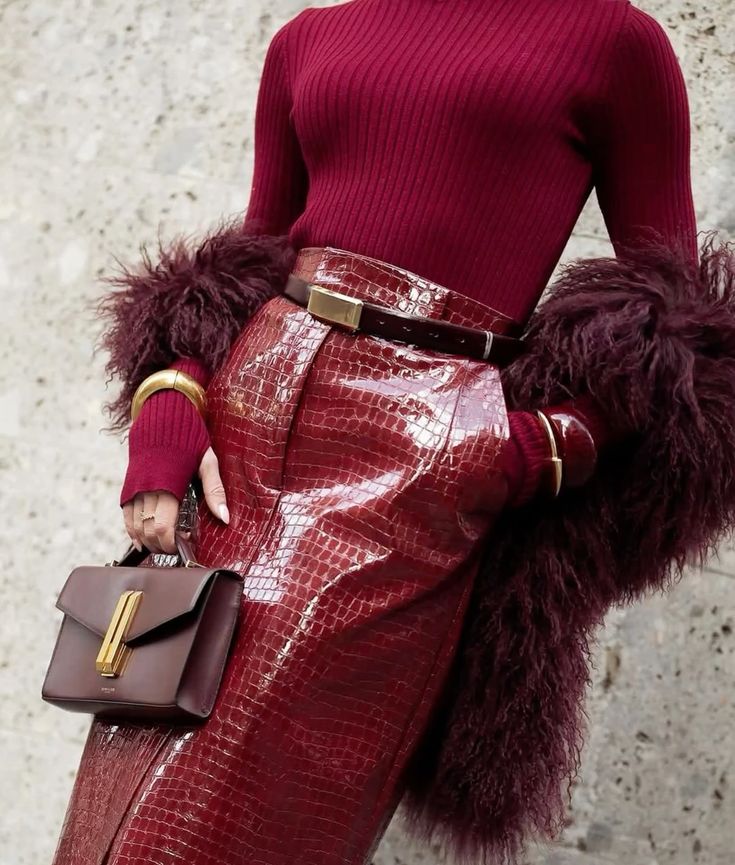 Fashionable woman in burgundy ensemble with textured skirt, sweater, fur coat, gold accessories, and handbag. Elegant style.