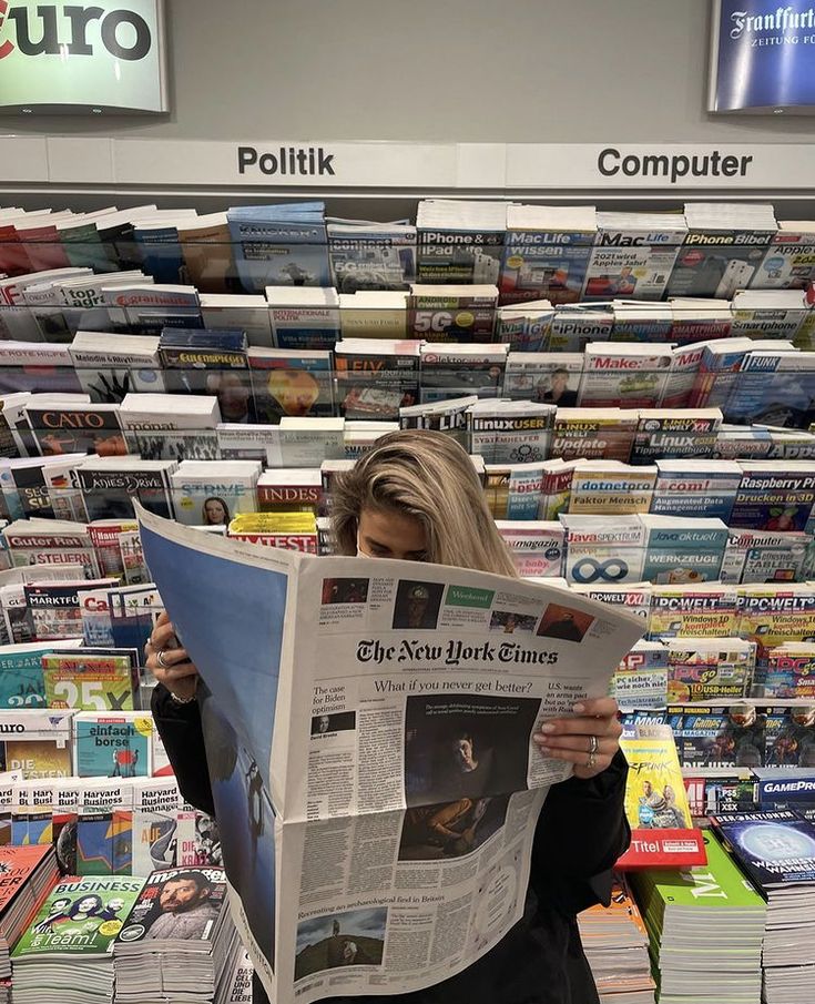 Woman reads the New York Times in front of a well-stocked magazine rack in a store.