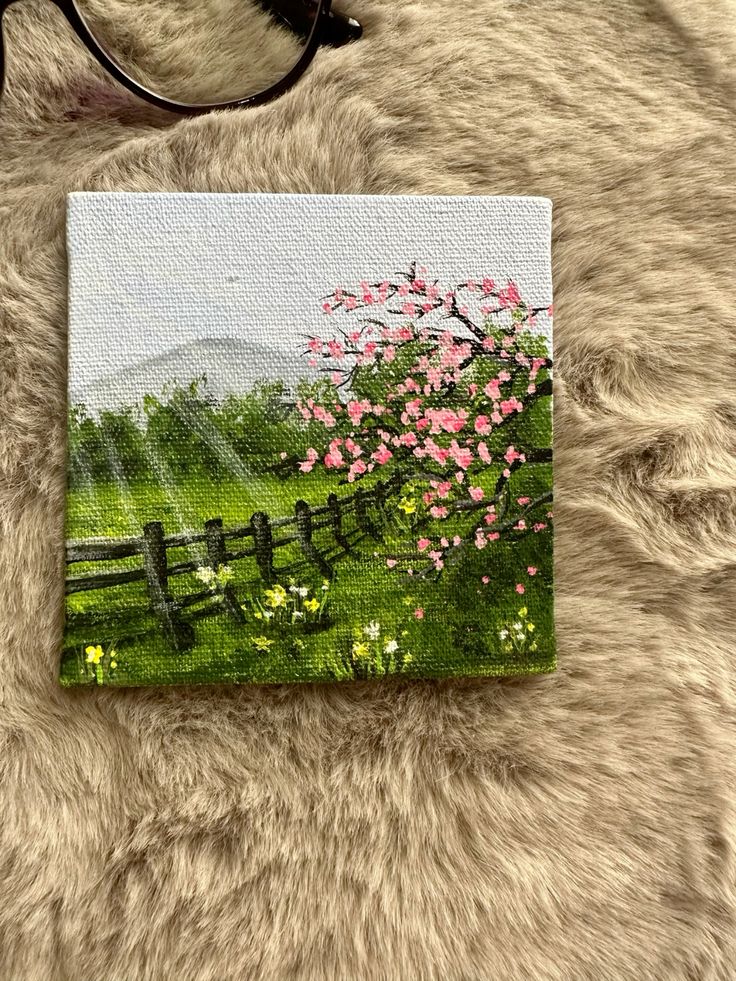 A small canvas painting of a serene spring landscape with pink blossoms, a fence, and grassy fields on a textured surface. | Sky Rye Design A small canvas painting of a serene spring landscape with pink blossoms, a fence, and grassy fields on a textured surface.
