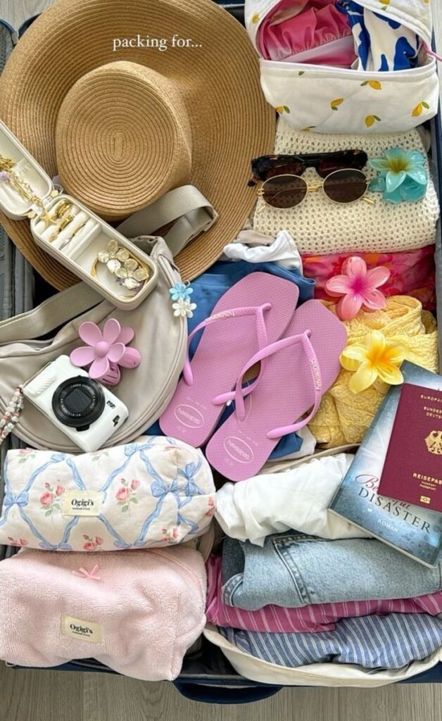 Open suitcase with beach hat, pink flip-flops, camera, passport, clothes, and accessories ready for summer vacation.