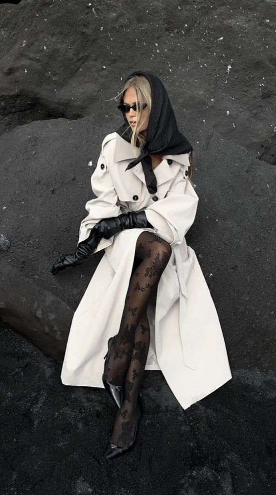 Fashionable woman in a trench coat, gloves, sunglasses, and patterned tights sits on dark rock background.