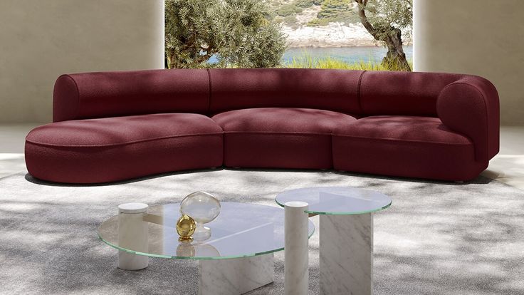 Curved burgundy sectional sofa with modern glass coffee tables in a spacious, light-filled room with a garden view.