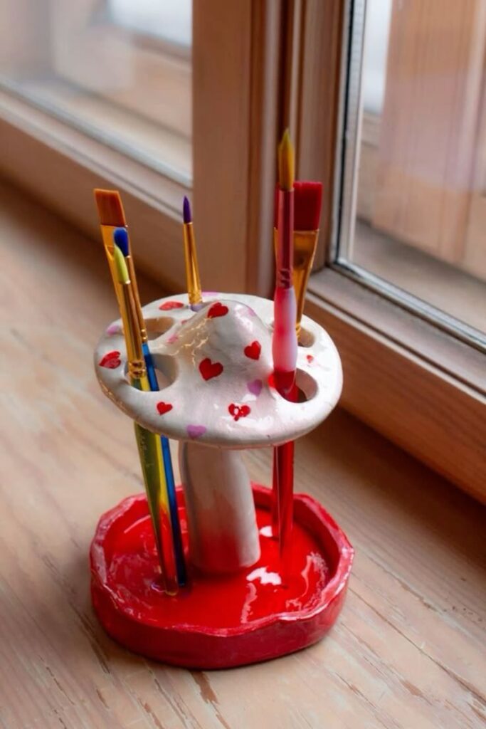 Mushroom-shaped paintbrush holder with colorful brushes, decorated with red hearts, placed by a wooden window.