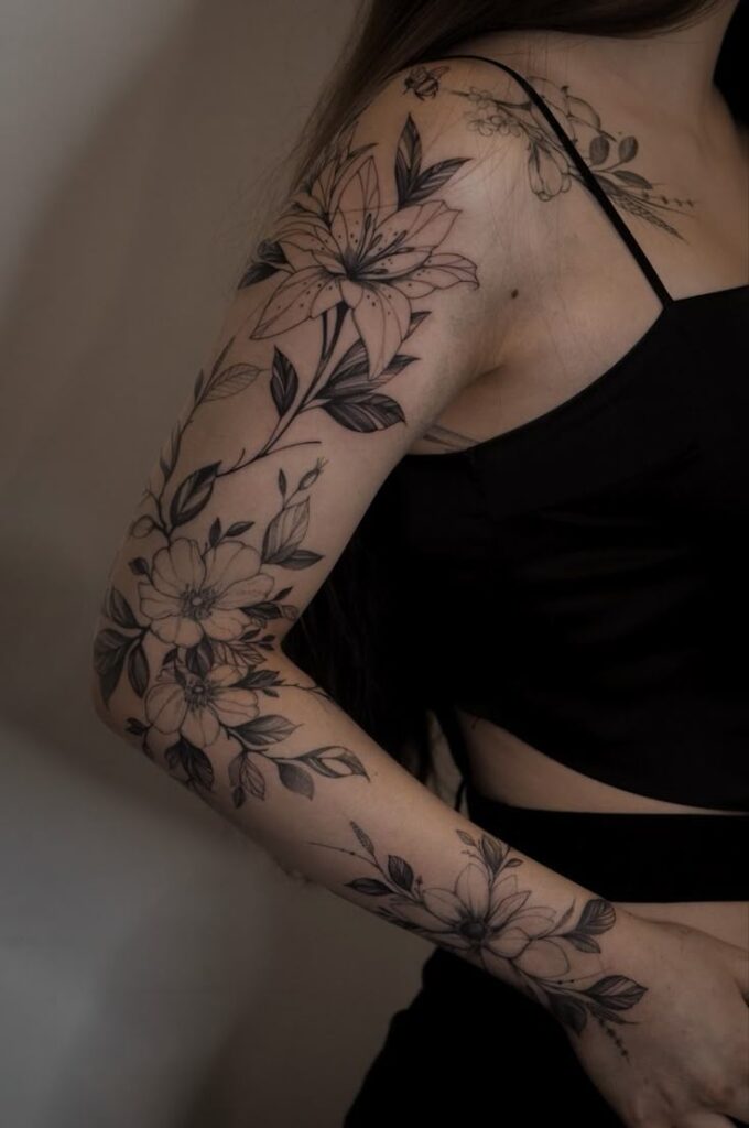 Woman's arm adorned with intricate floral tattoos in black ink, wearing a black sleeveless top.