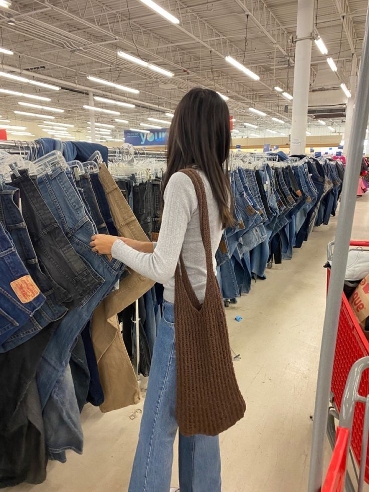 Person shopping for jeans in a store aisle, wearing a brown bag and light sweater, surrounded by hanging denim.