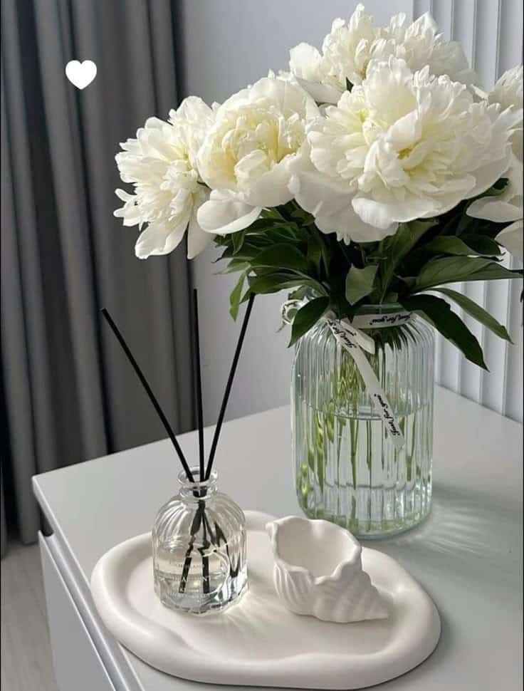 White peonies in a glass vase on a table with a reed diffuser and decorative dish.