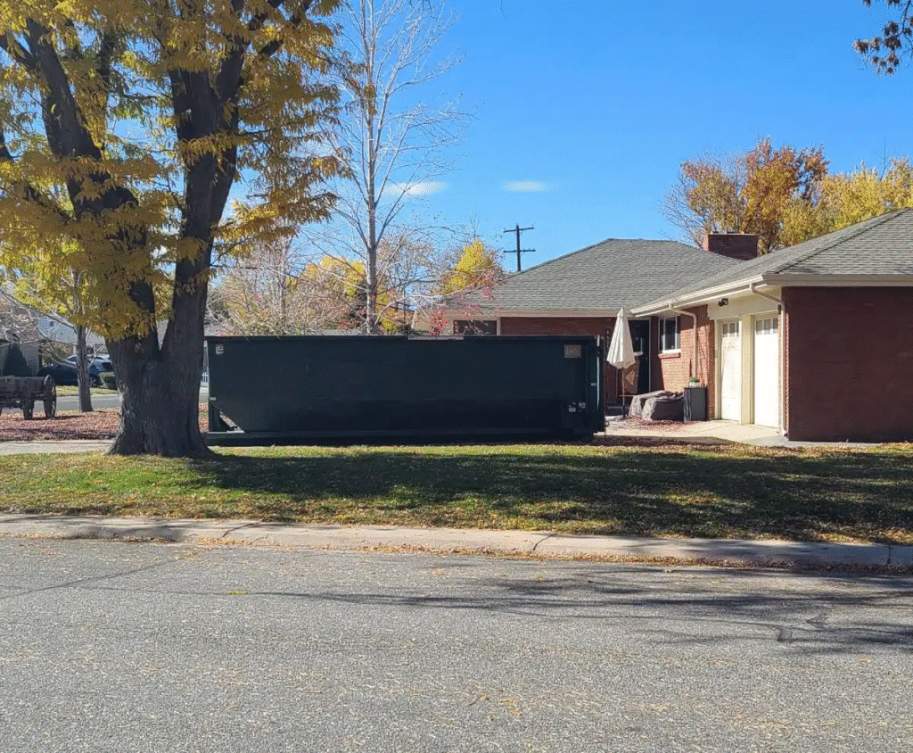 Dumpster in front of suburban house during autumn, surrounded by trees with yellow leaves.