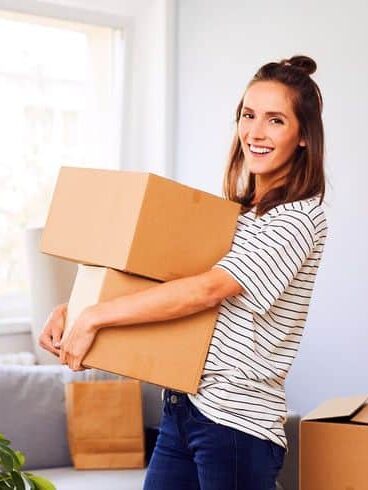 Smiling woman carrying moving boxes in a bright room with plants and sunlight through the window.