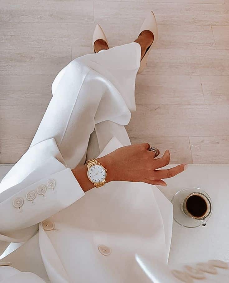 Elegant woman in white suit with a gold watch, holding coffee, wearing beige heels, sitting stylishly on beige flooring.