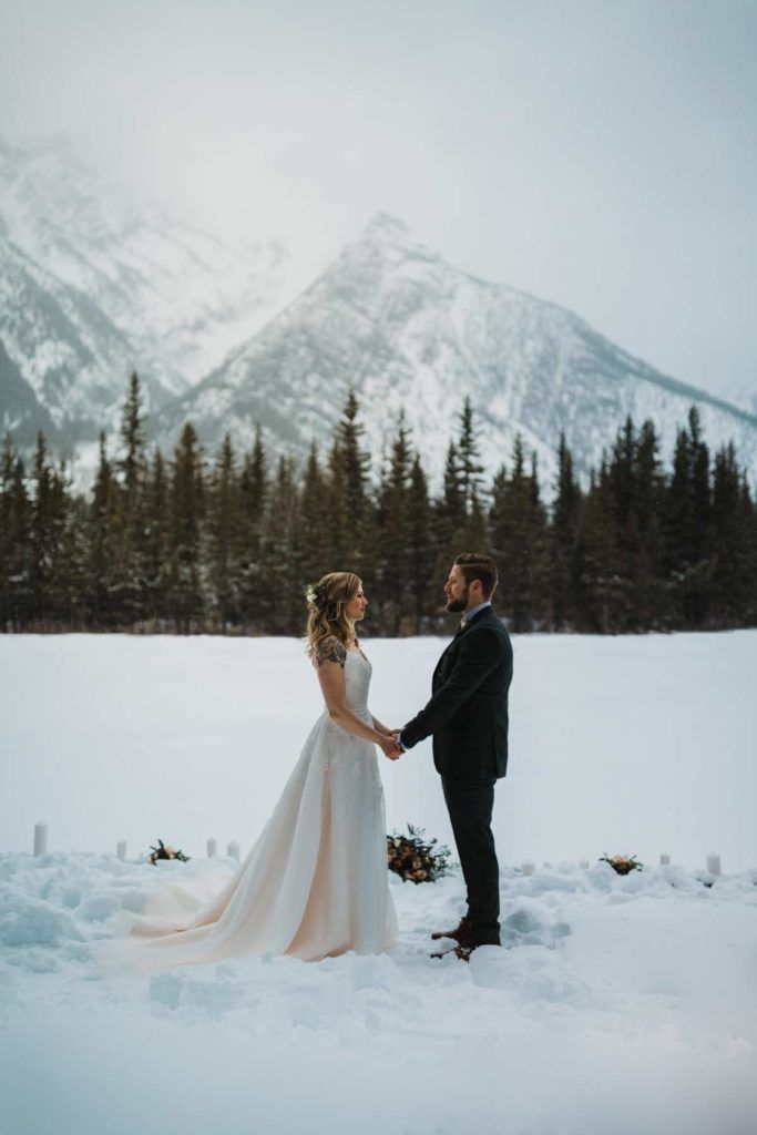 Bride and groom holding hands in snowy mountain landscape, surrounded by trees.