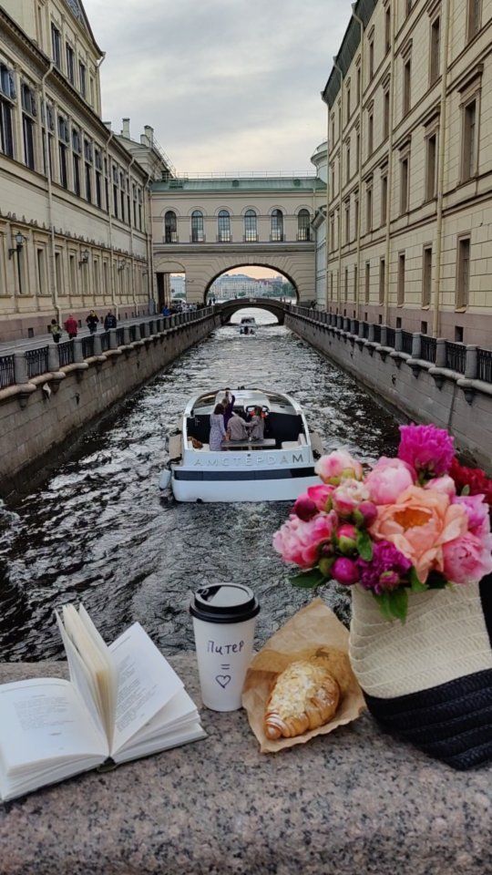 Canal boat tour with book, coffee, croissant, and flowers in a scenic city setting.