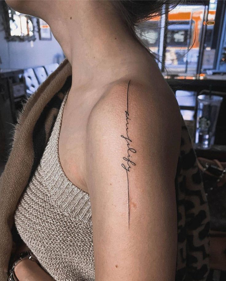 Minimalist cursive arm tattoo on person wearing a knitted tank top in a cozy café setting.