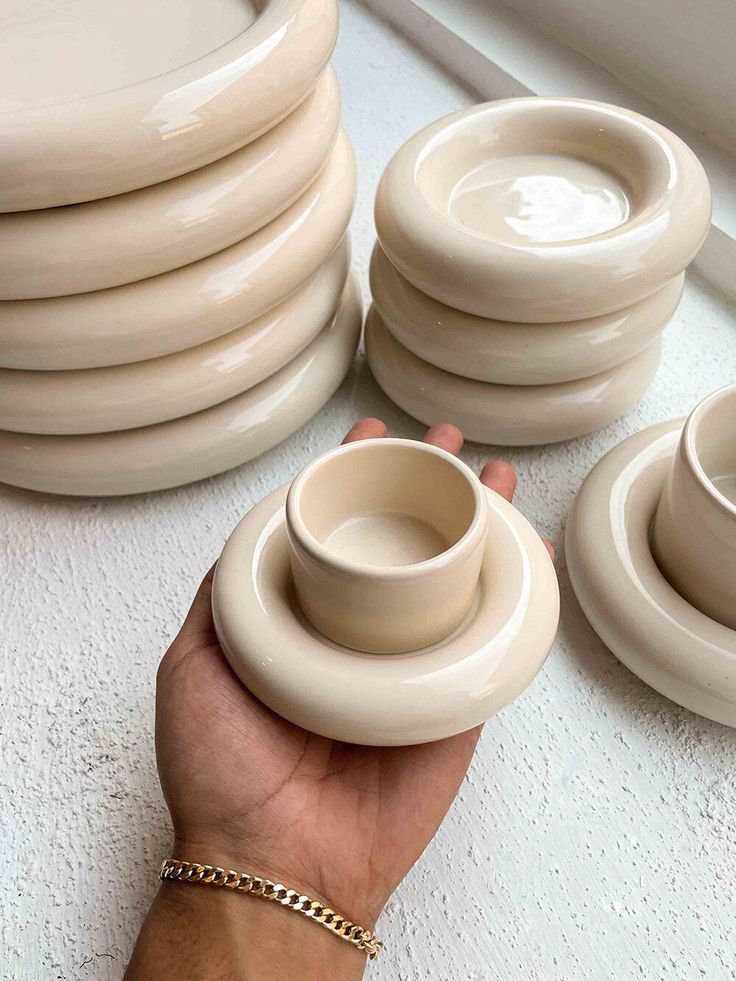 Hand holding a cream ceramic cup and saucer set, with stacks of similar dishes in the background on a white surface.