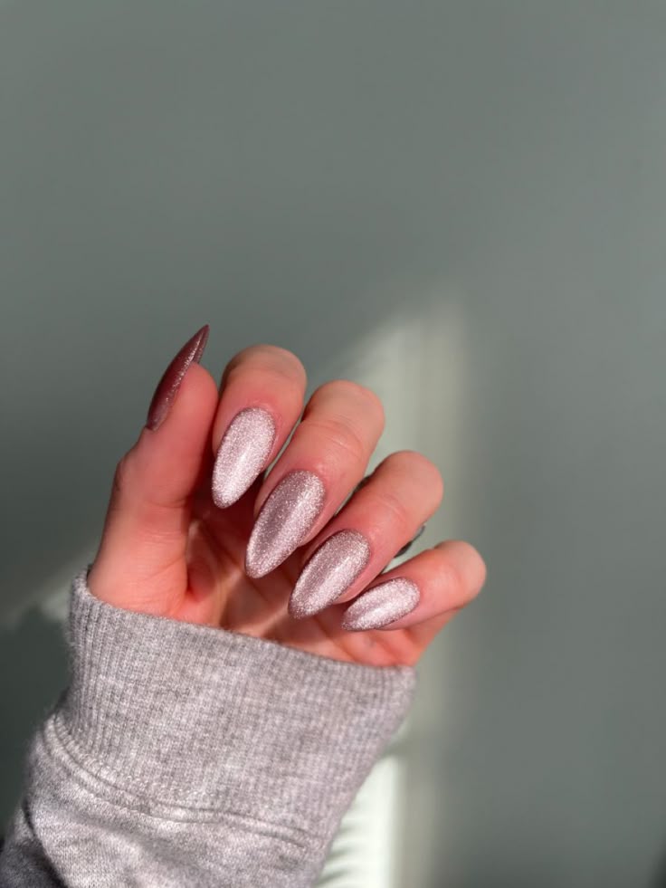 Hand with long, almond-shaped nails painted in glittery pink polish, sweater sleeve visible, against a soft background.