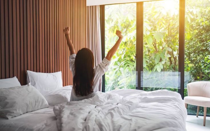 Woman stretching on bed in bright room with large window and greenery outside, embracing a refreshing morning start.