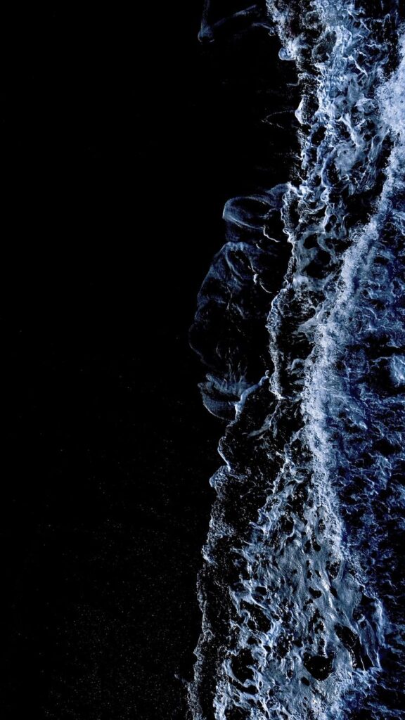 Aerial view of white ocean waves crashing onto a dark sandy beach at night.