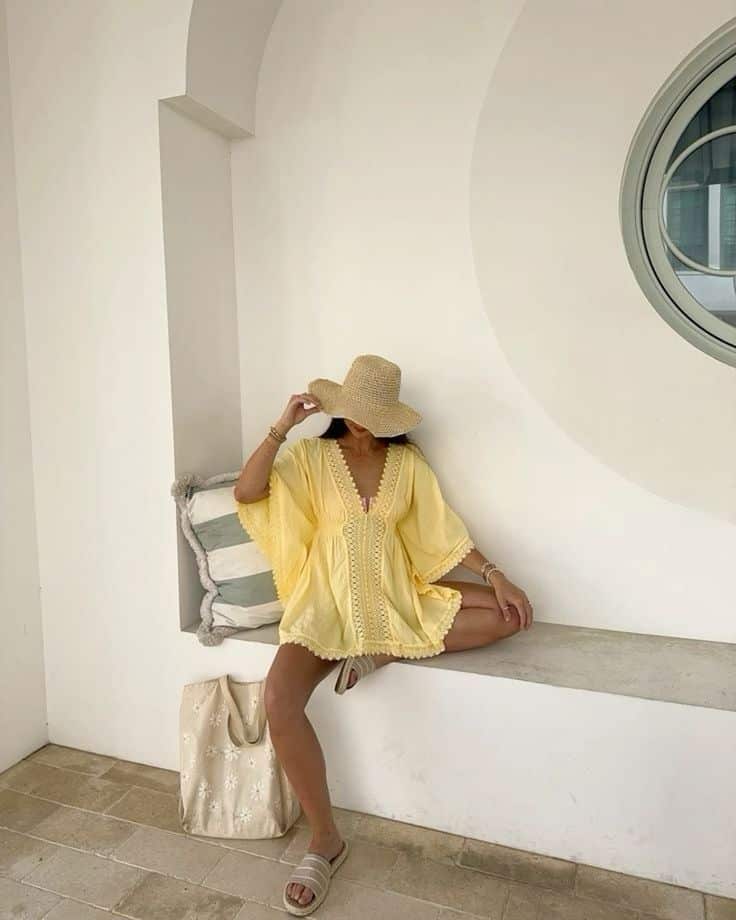 Woman in yellow dress and straw hat poses on a white bench beside a large tote bag, against a stylish architectural backdrop.