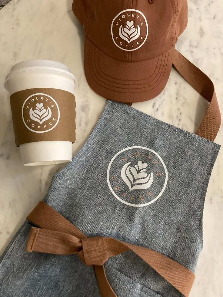 16ca4d77-defc-4676-83d1-348449478115 Coffee cup, cap, and apron with Violet's Coffee logo on a marble surface. | Sky Rye Design Coffee cup, cap, and apron with Violet's Coffee logo on a marble surface.