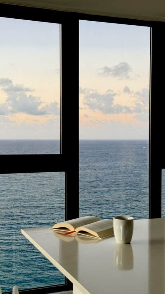Cozy ocean view with open book and coffee at sunrise through large window.