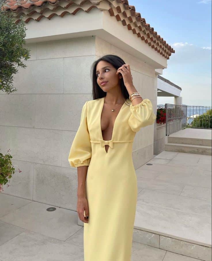 Woman in elegant yellow dress poses outdoors near a modern building on a sunny day.